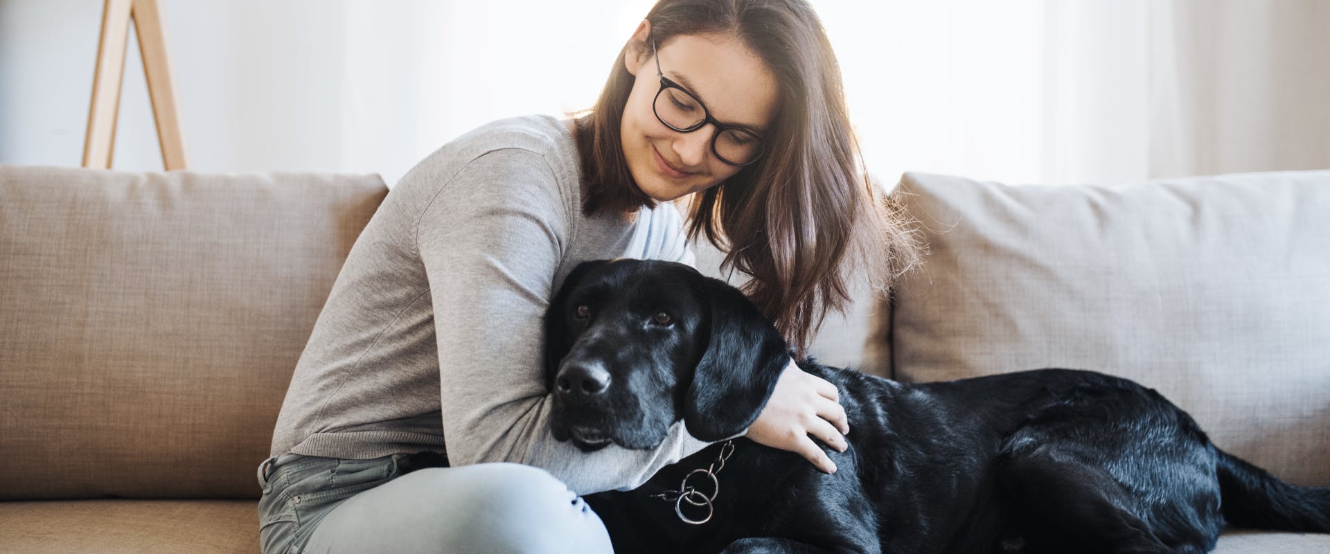 a smiling pet sitter wearing glasses sitting on a gray couch cuddling a black Labrador