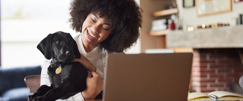 a woman sat smiling in front of a laptop while cuddling and smiling at a black puppy
