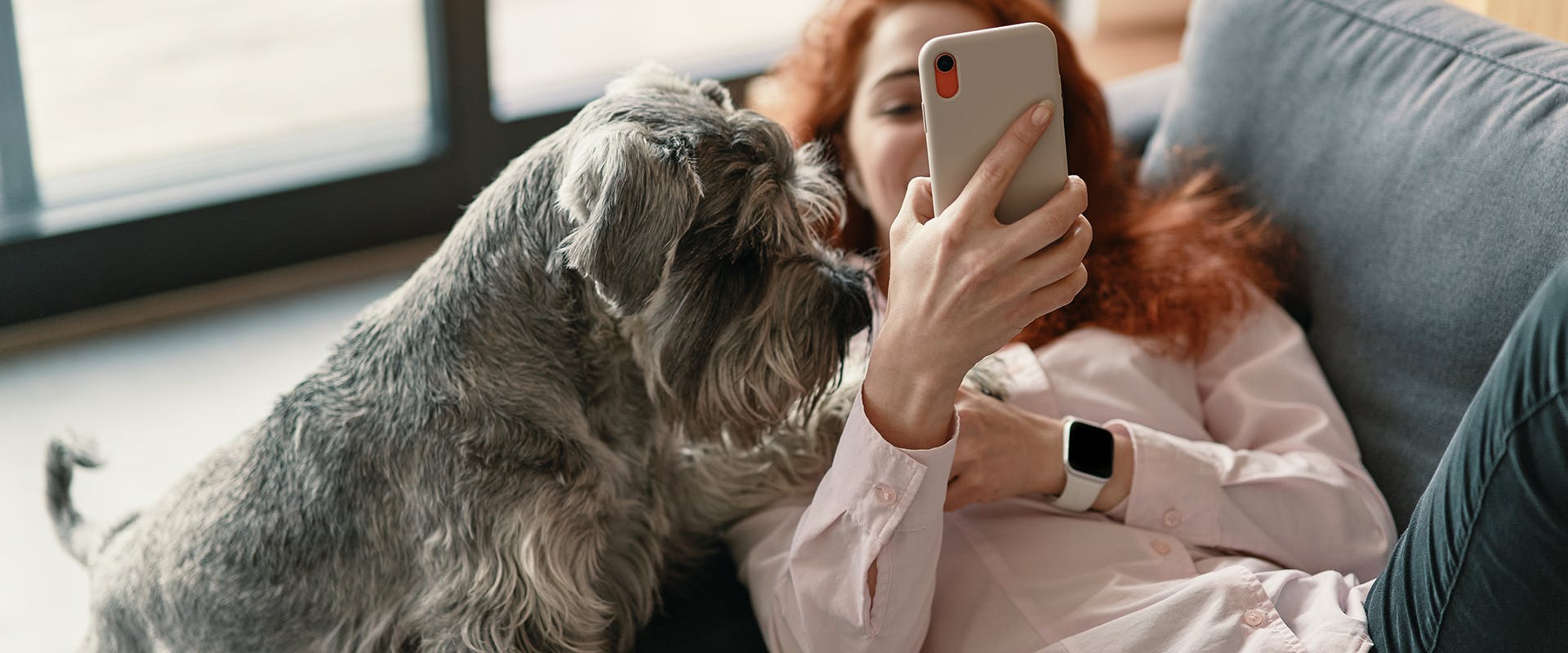 A woman laying on a sofa looking at her phone, a small dog at her side