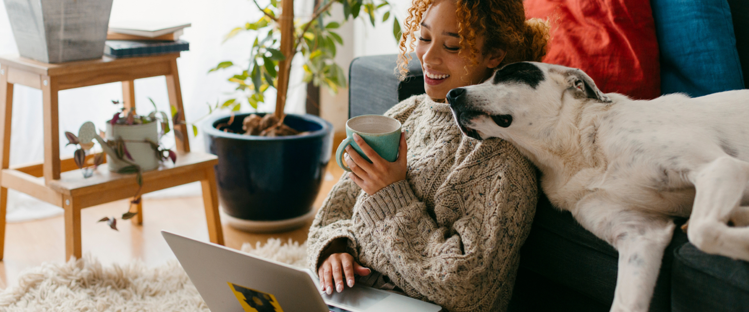 A woman sitting on the floor on her laptop, a dog sleeping on the sofa behind her with its head resting on the woman's shoulder
