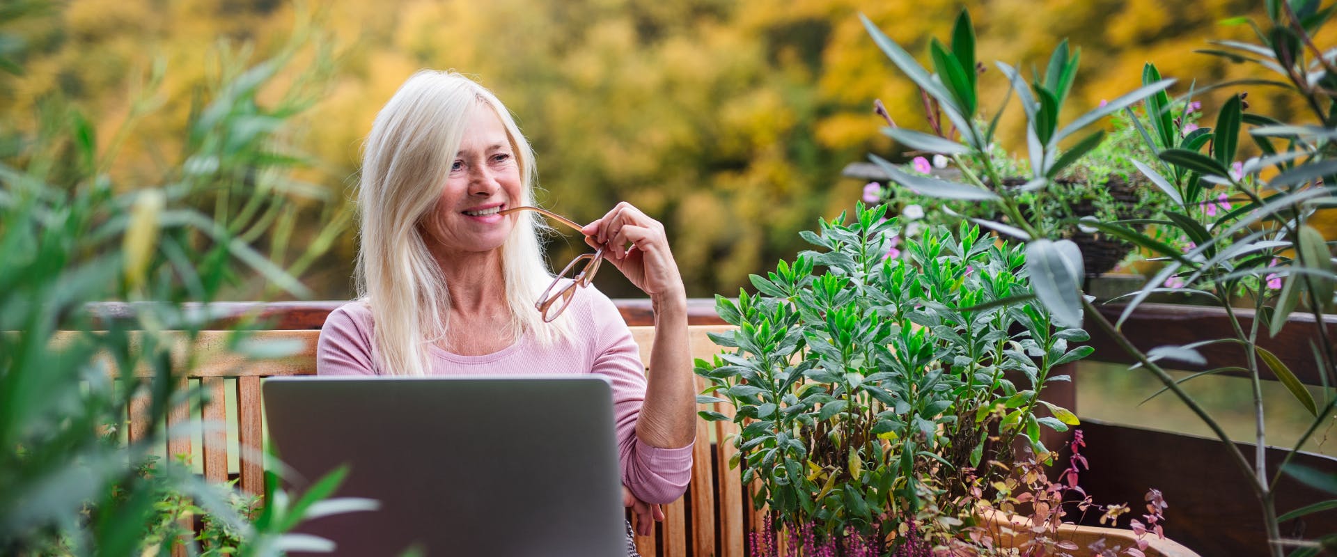 a woman sat on a wooden bench surrounded by shrubs with an open laptop on her lap in a rural area