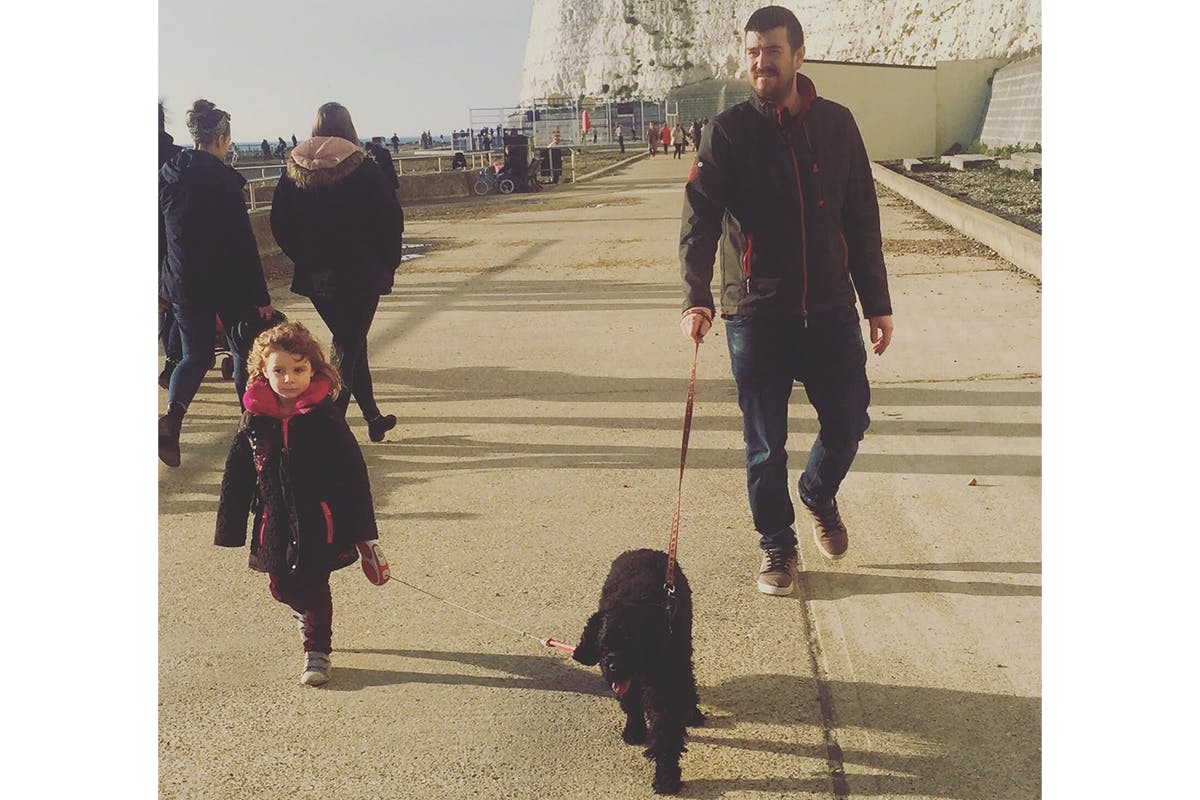 A man, a child, and a black dog walking under Brighton cliffs