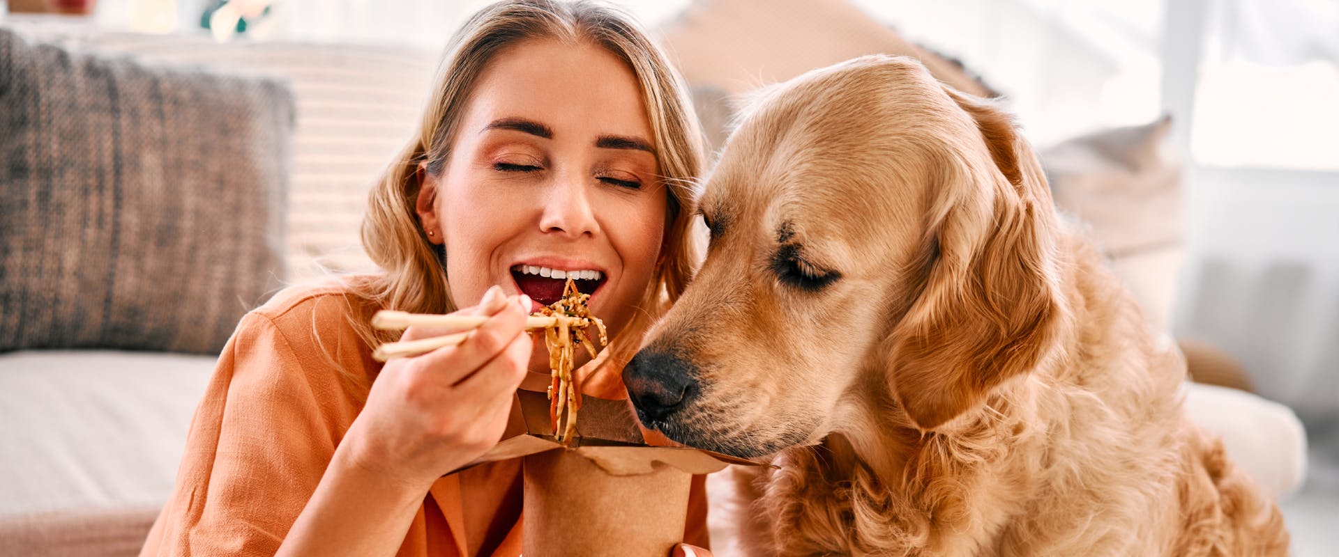 a woman using chopsticks to eat noodles with a curious golden retriever right next to her face sniffing the noodles