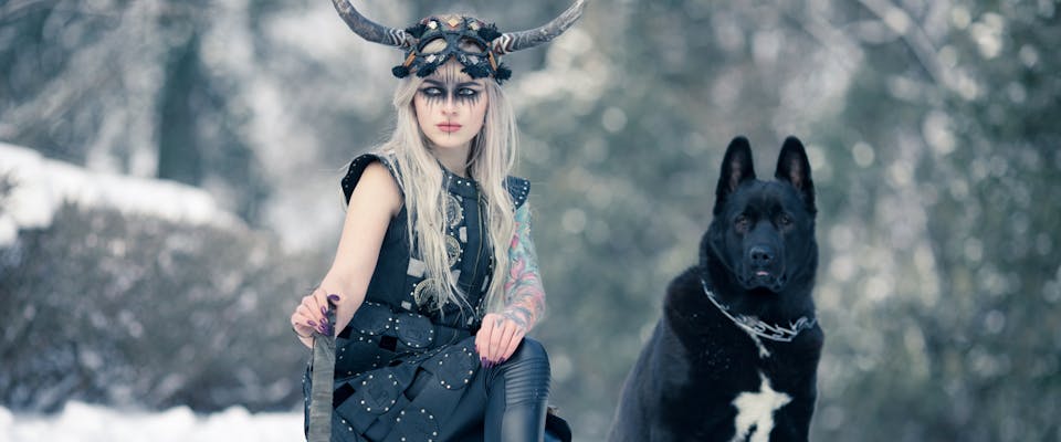 woman dressed as a viking next to a black and white dog in a snowy forest