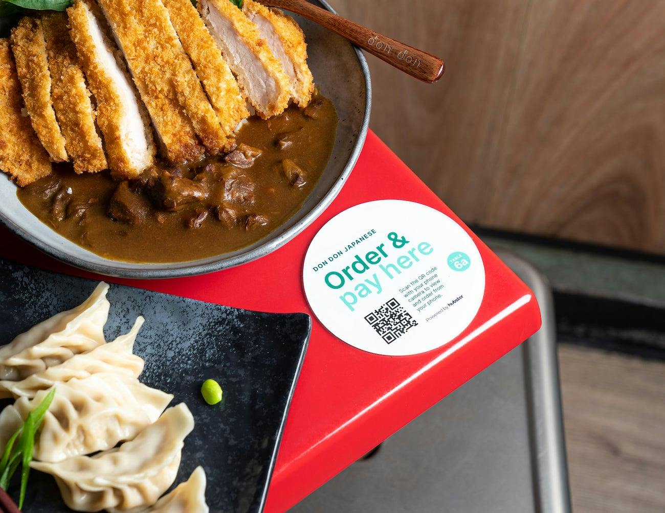 Image of a QR code ordering coaster on the corner of a table with curry rice and dumplings.