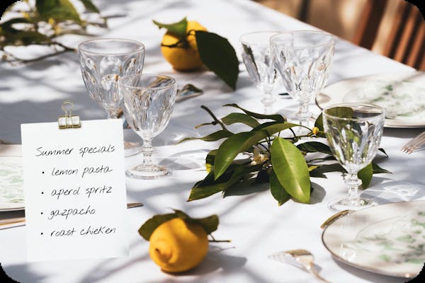 Image of a sunny table setting with a note of summer menu specials: lemon pasta, aperol spritz, gazpacho and roast chicken
