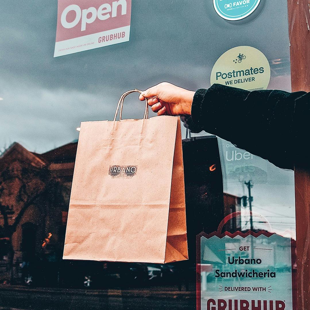 Image of Urbano Food Group employee holding out a delivery bag in front of their restaurant's window which has clings for Postmates, Grubhub and more