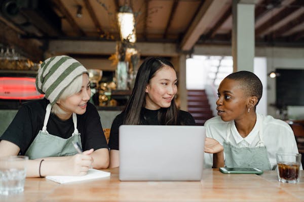 Image of three women sitting in front of a laptop in a cafe