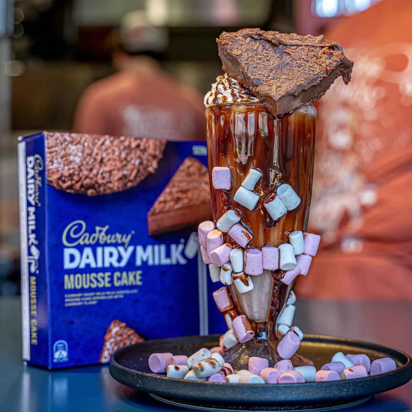 Image of a glass of marshmallow chocolate milkshake, with a box of Cadbury dairy milk cake in the background