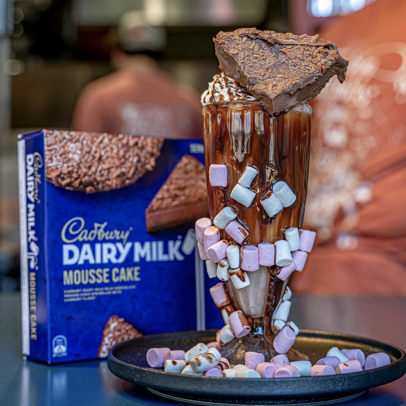 Image of a glass of marshmallow chocolate milkshake, with a box of Cadbury dairy milk cake in the background