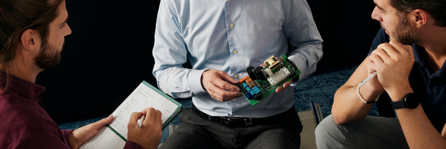 Three men in business attire examine and discuss an electronic circuit board during what appears to be a technical workshop or meeting.