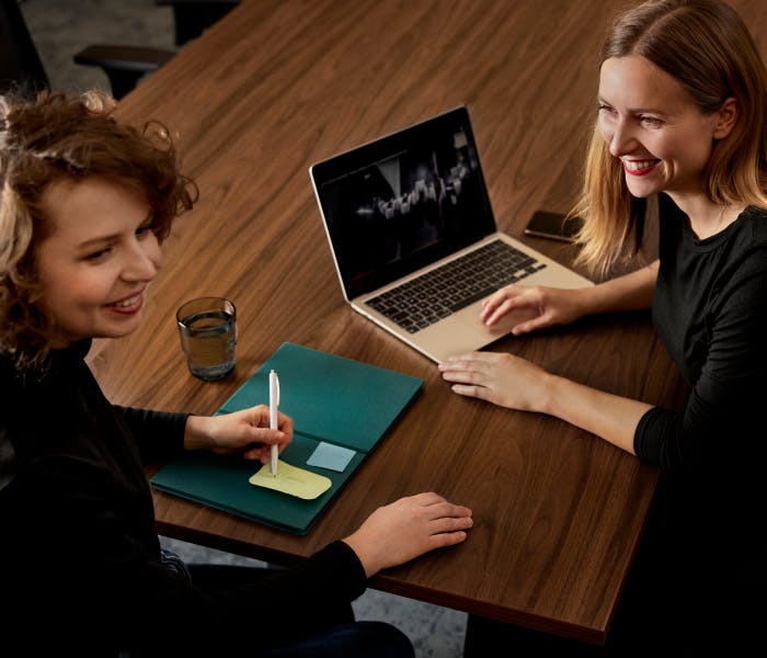 Two smiling women sit at a wooden desk, one using a laptop displaying a graph while the other takes notes, suggesting a personalized guidance or mentoring session.