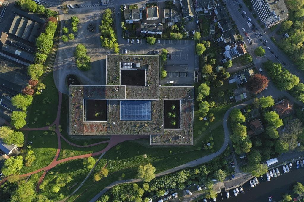Aerial top-down view of a Dräger building with a green roof, solar panels, and inner courtyards, surrounded by trees, walking paths, and a nearby canal with boats—showcasing the company’s sustainable architecture and integration with the natural environment.