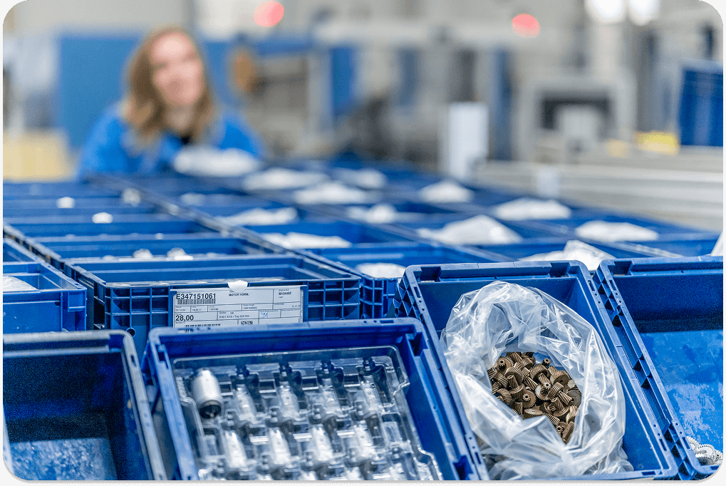 Close-up of labeled blue crates filled with industrial components in a production warehouse, with a blurred worker in the background
