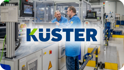 Küster logo in front of the two engineers in blue lab coats discussing a component at an automated workstation in a modern manufacturing facility.