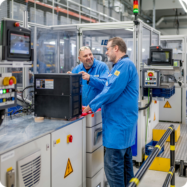 Two engineers in blue lab coats discussing a component at an automated workstation in a modern manufacturing facility.
