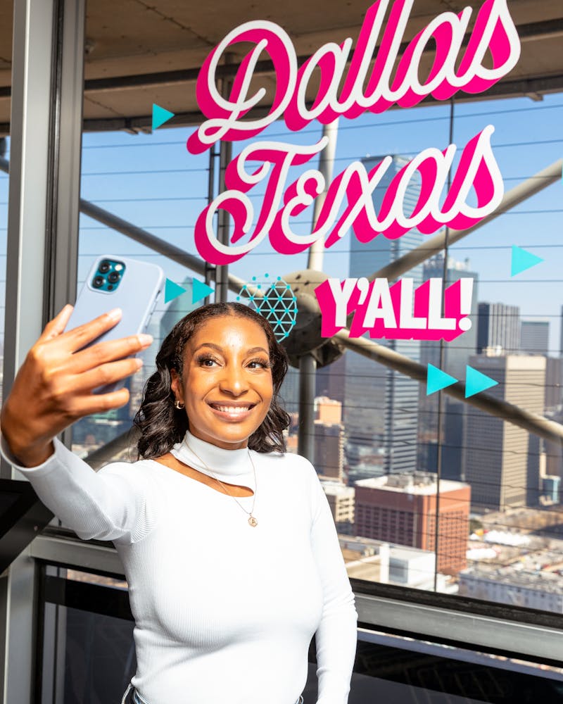 woman taking a selfie at Reunion Tower in front of a window decal that reads "Dallas Texas, Y'all!"