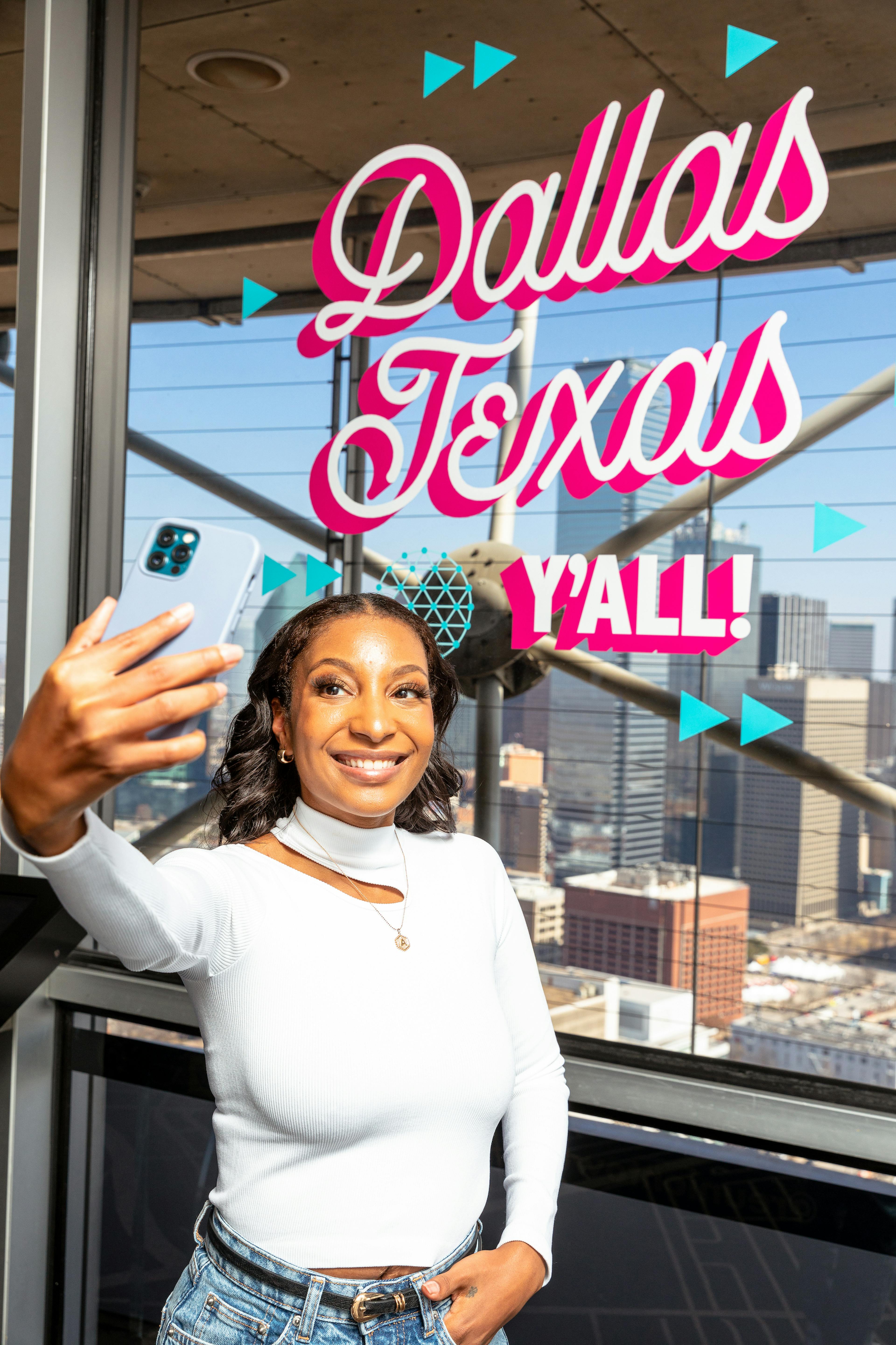 woman taking a selfie at Reunion Tower in front of a window decal that reads "Dallas Texas, Y'all!"