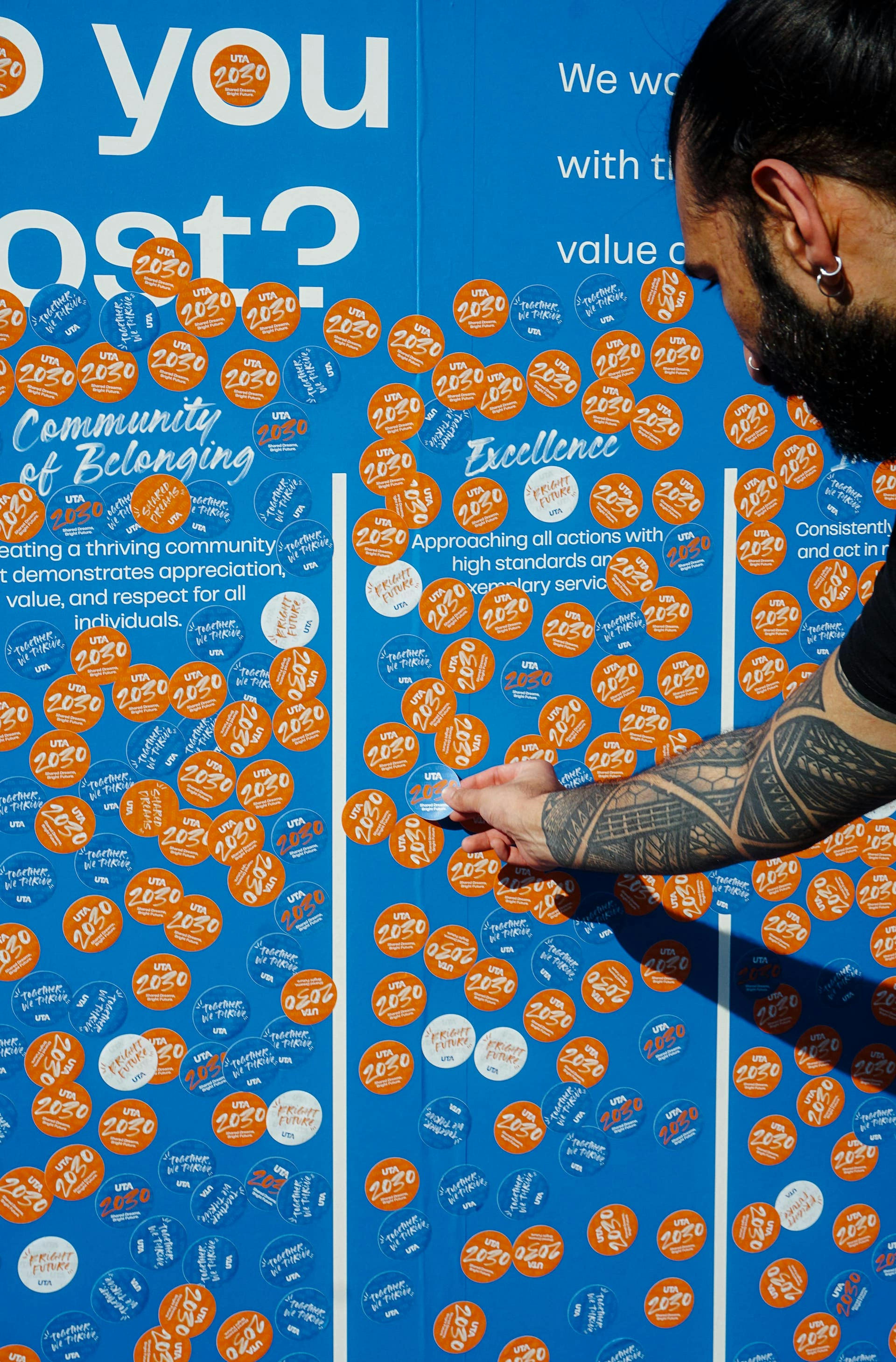 A person is placing an orange "UTA 2030" sticker on a blue wall filled with similar stickers, promoting community and excellence.