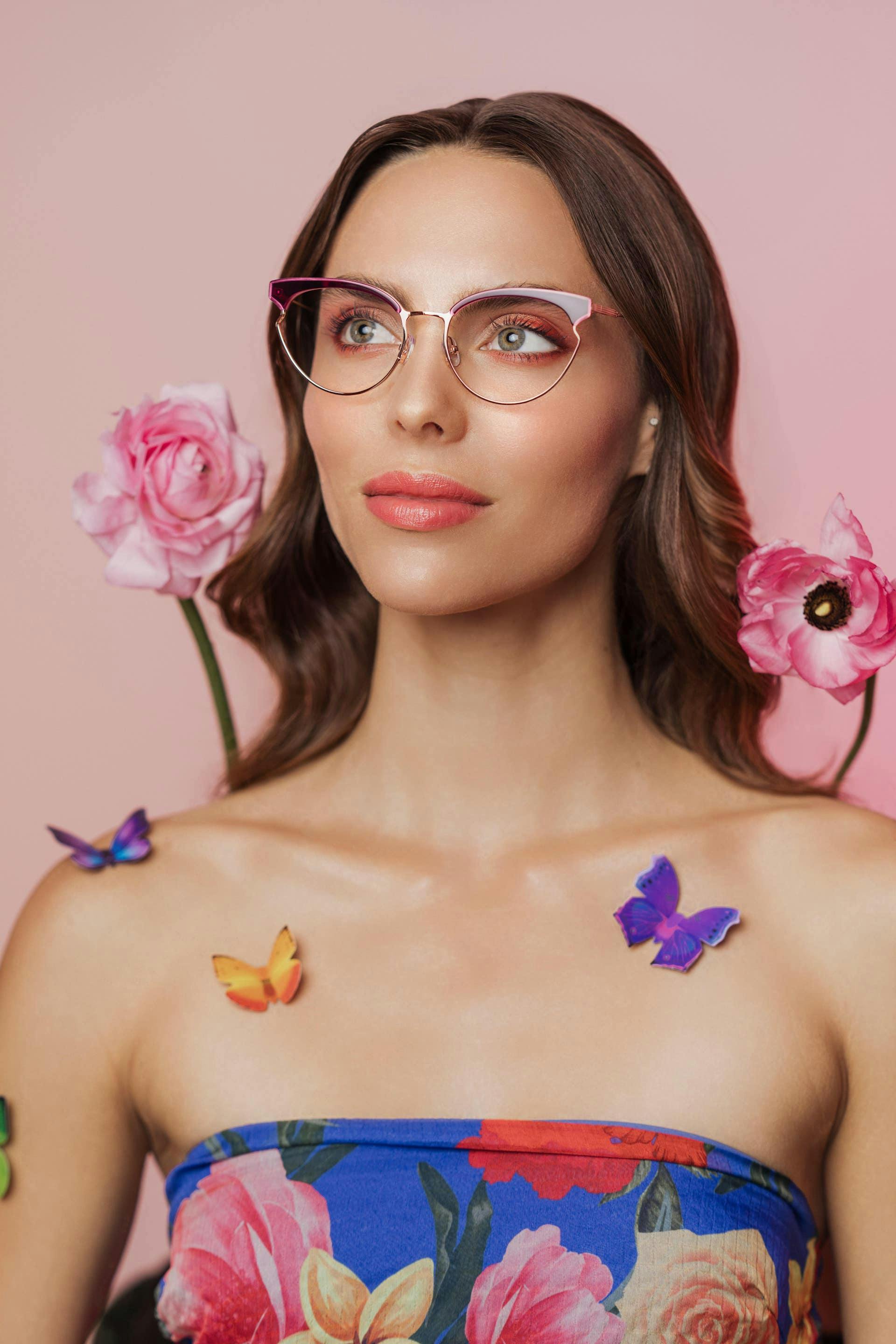 A woman modeling a pair of RACHEL Rachel Roy glasses, wearing a floral outfit on pink background.