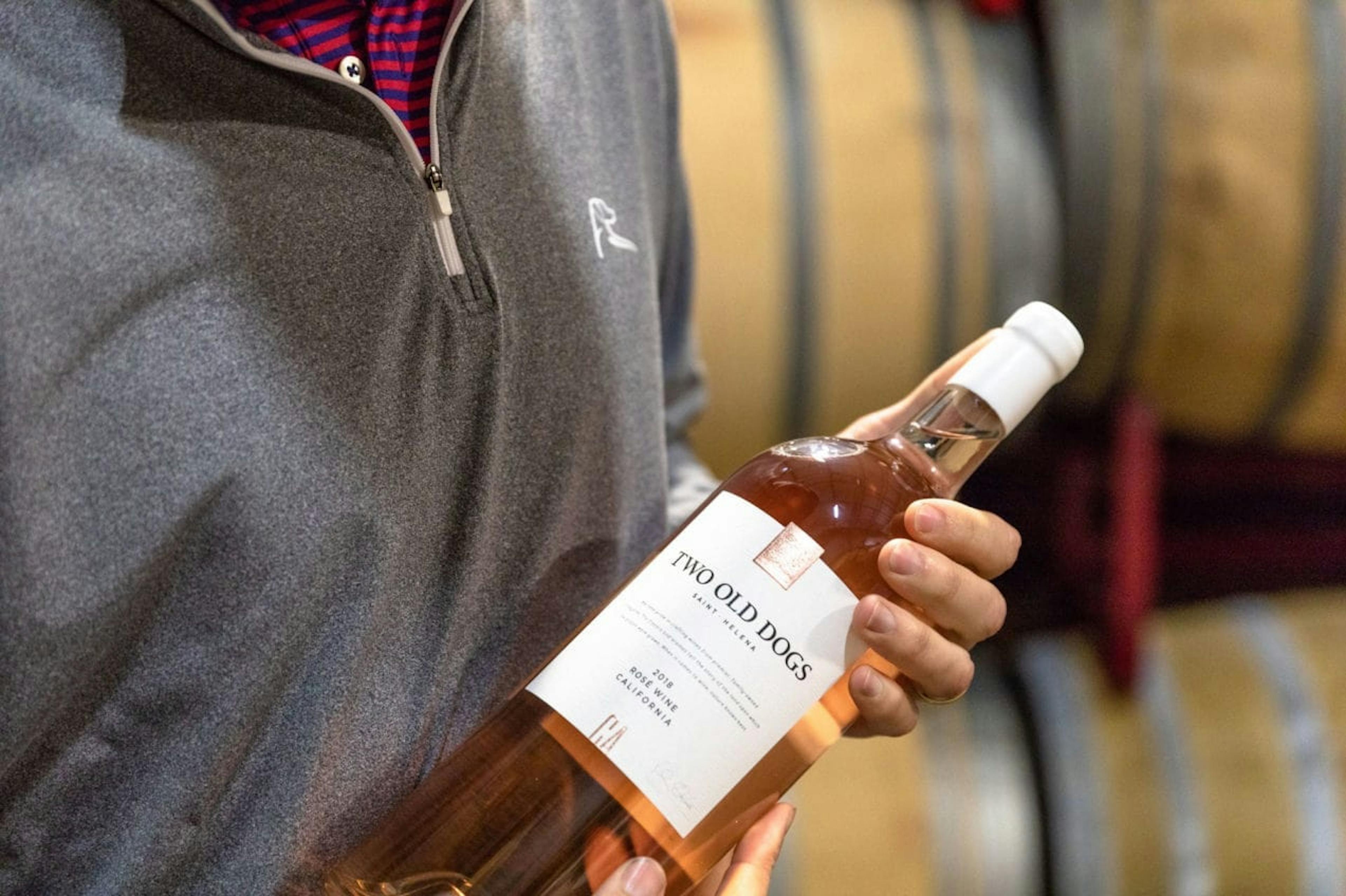 Hand holding a bottle of Two Old Dogs wine surrounded by wine barrels