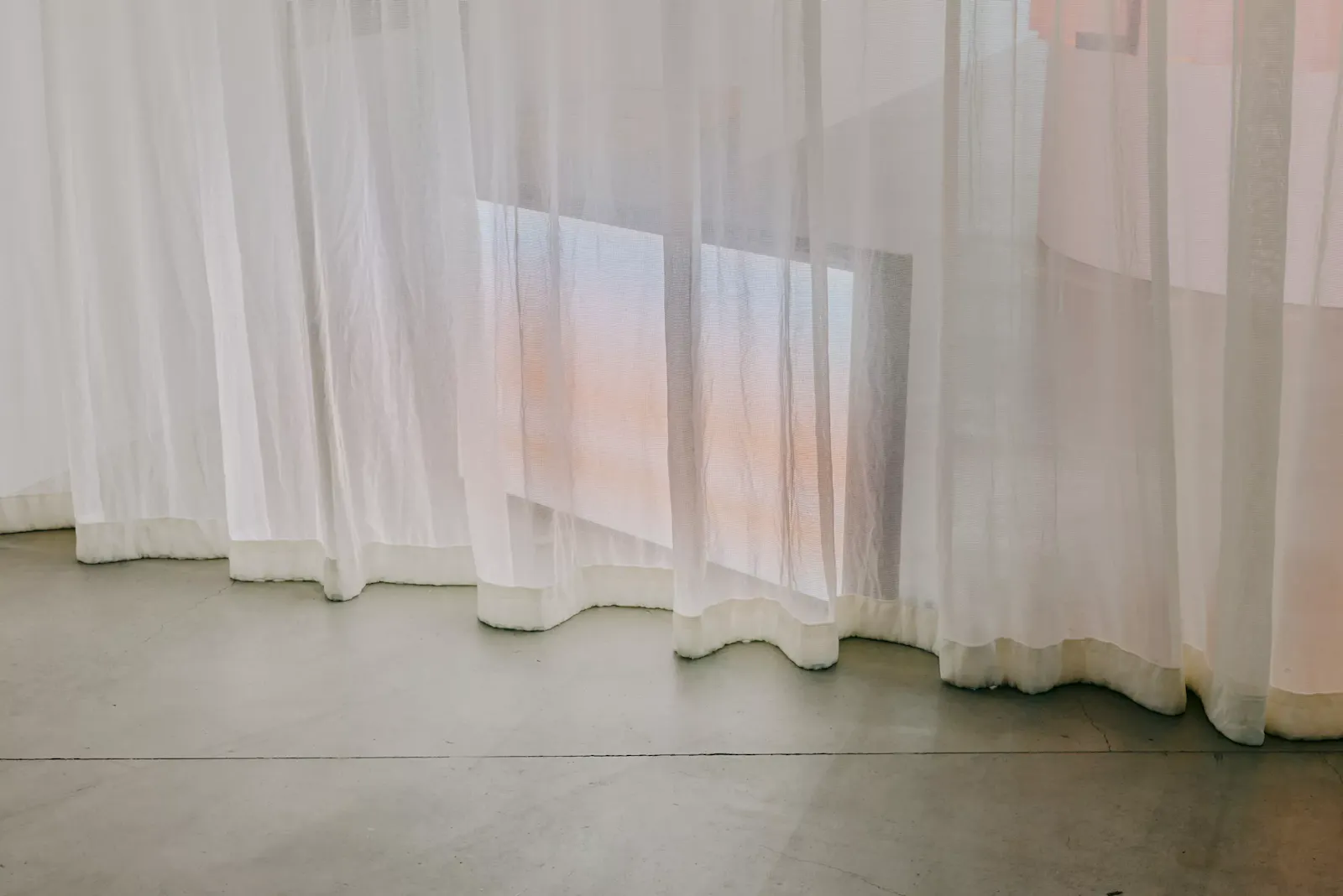 Gallery installation view of The Mourning After at RMIT Design Hub. Sheer translucent curtains hang across a quiet concrete-floored room, washed in soft pastel pink and blue light, creating a contemplative, ethereal space. U-P(co)2010
