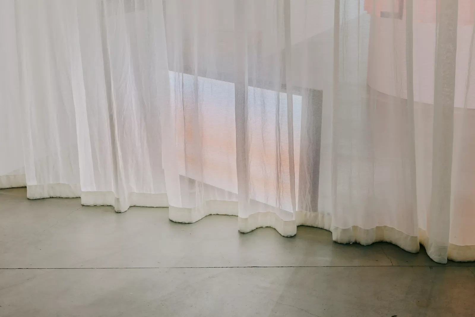 Gallery installation view of The Mourning After at RMIT Design Hub. Sheer translucent curtains hang across a quiet concrete-floored room, washed in soft pastel pink and blue light, creating a contemplative, ethereal space. U-P(co)2010