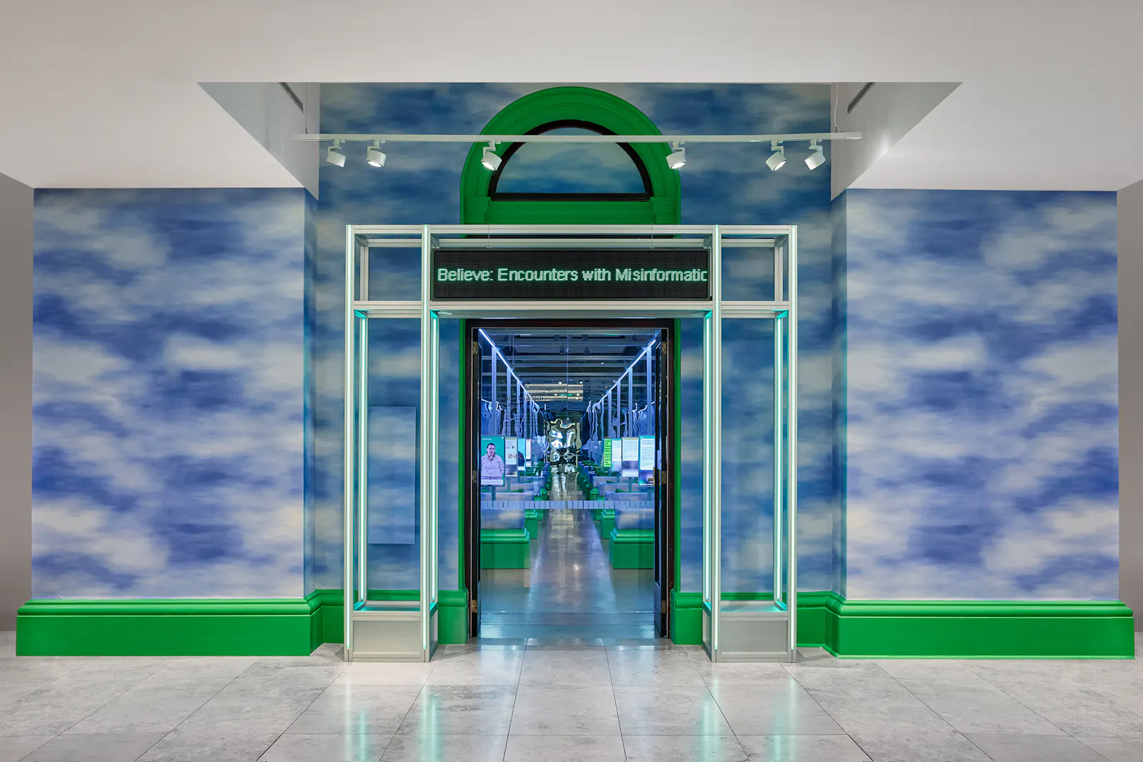 Exhibition entrance for Make Believe: Encounters with Misinformation at State Library Victoria. A bright green archway frames a doorway leading into a long mirrored corridor, with walls clad in soft blue-and-white cloud imagery. A small overhead sign reads "Believe: Encounters with Misinformation." U-P(co)2010