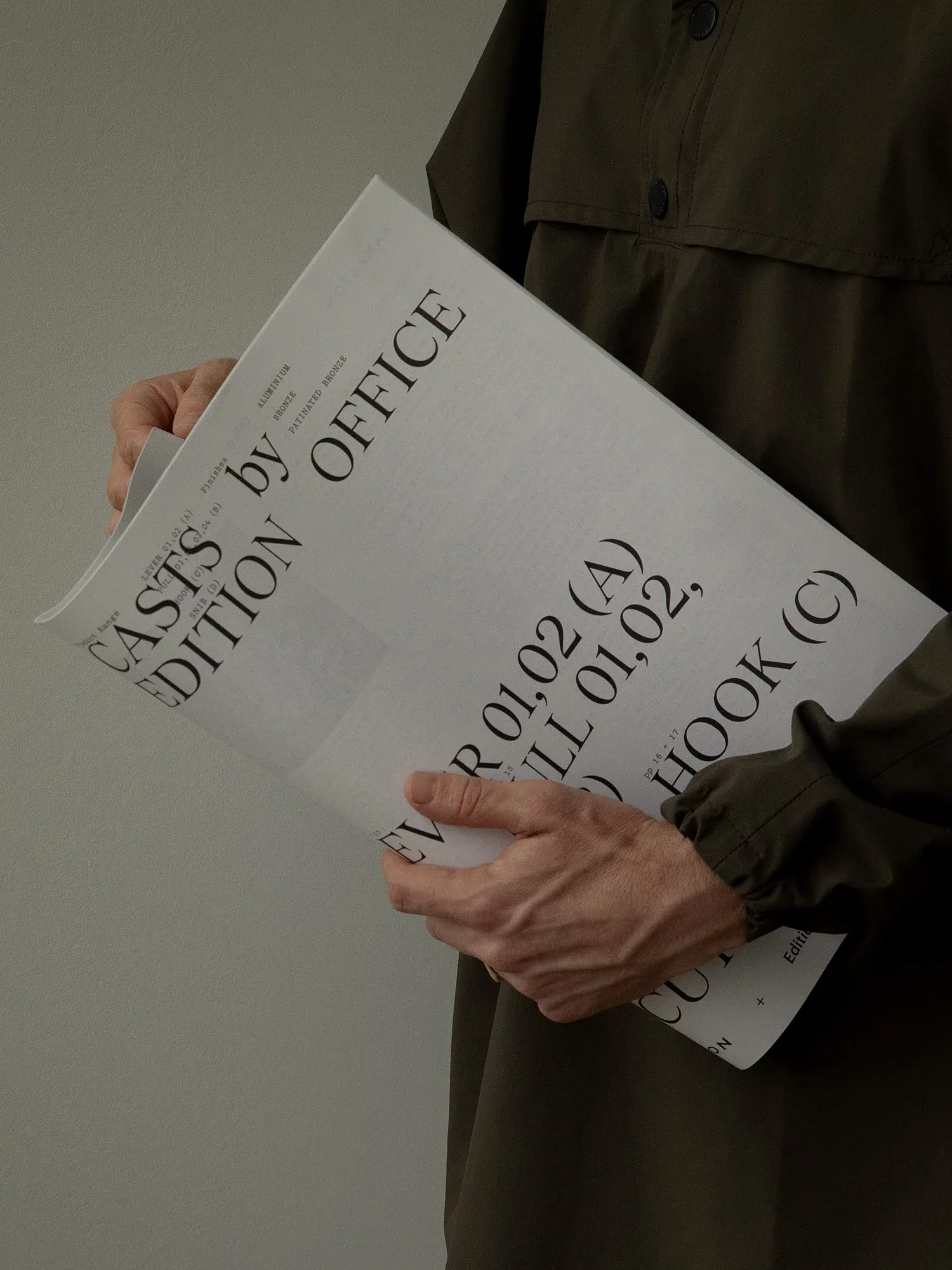 Creative direction for the Casts door handle collection, a Bankston x Edition Office collaboration. A person in a dark jacket holds a large folded printed poster or specification sheet set in elegant serif typography, with fragments reading "Casts by Edition Office" and product references. U-P(co)2010