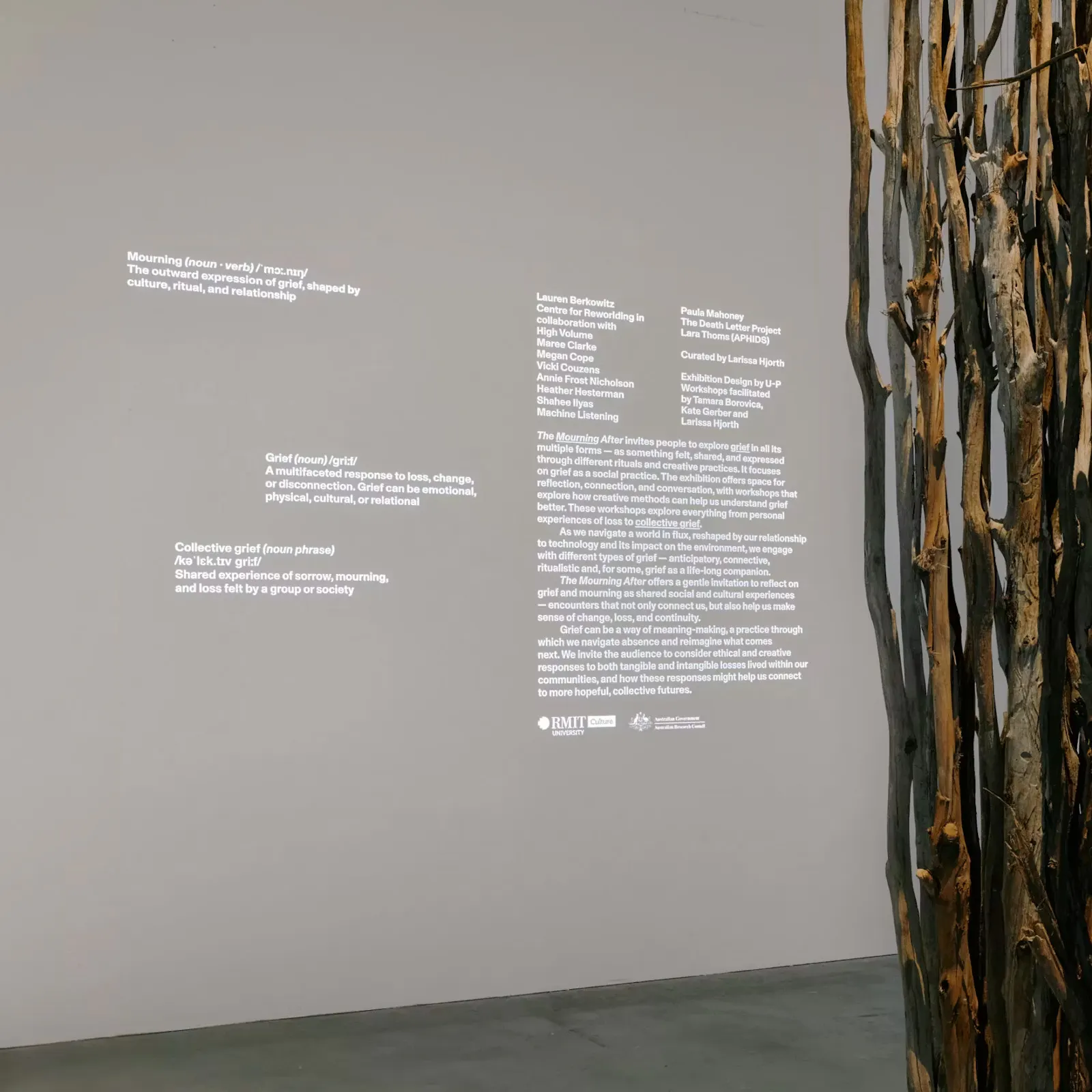 Installation view of The Mourning After at RMIT Design Hub. Blocks of small white exhibition text are projected or applied across a pale gallery wall, beside a vertical sculptural element made of bundled branches or driftwood. U-P(co)2010