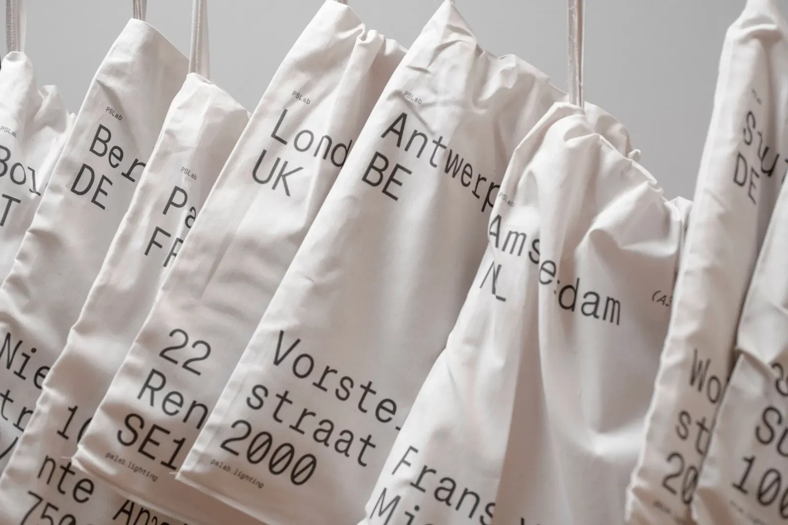 Creative direction for lighting company PSLab. A row of natural canvas drawstring bags hangs in a line, each stamped with hand-drawn-style addresses for international cities including London UK, Antwerp BE and Amsterdam NL. U-P(co)2010