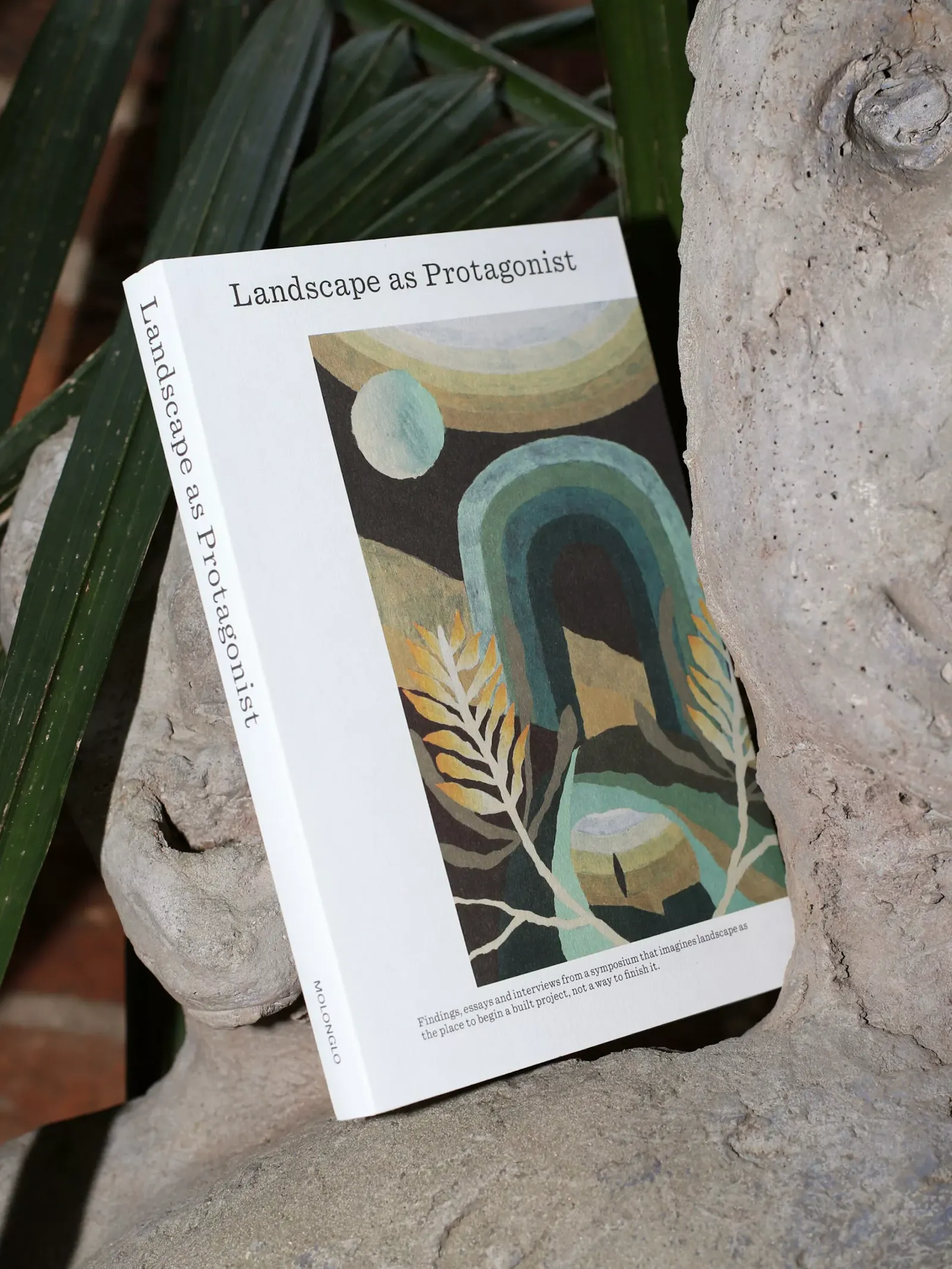 The Landscape as Protagonist publication for Molonglo. A white softcover book stands upright against pale stone and green foliage, its cover showing an abstract painterly composition of layered arches, leaves and organic shapes in muted greens, blues and ochre. U-P(co)2010