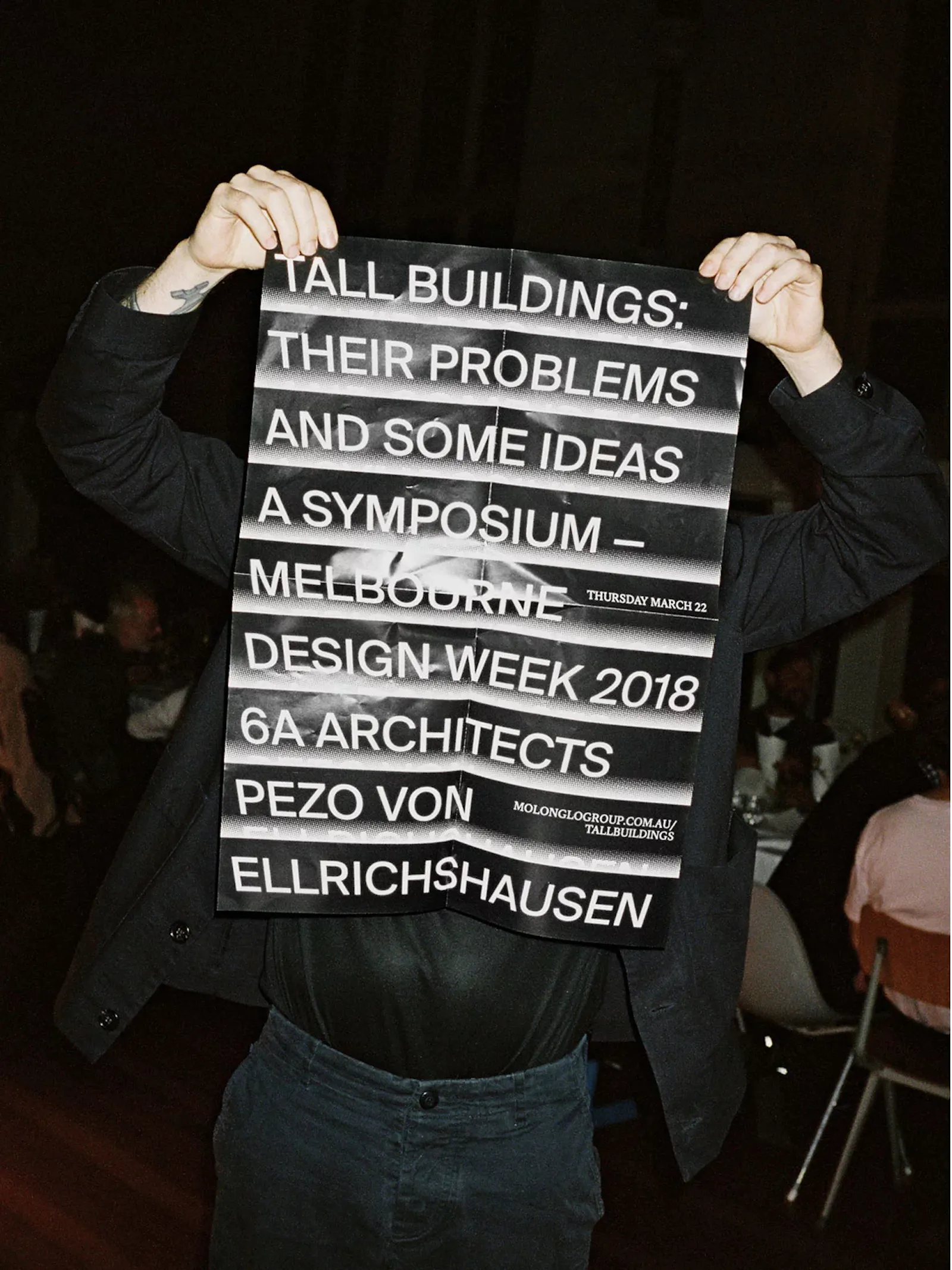 Identity for the Tall Buildings symposium at Molonglo. A person dressed in black holds a folded black-and-white poster across their face and torso, the bold condensed type reading "Tall Buildings: Their Problems and Some Ideas — A Symposium, Melbourne Design Week 2018, 6a Architects, Pezo von Ellrichshausen." U-P(co)2010