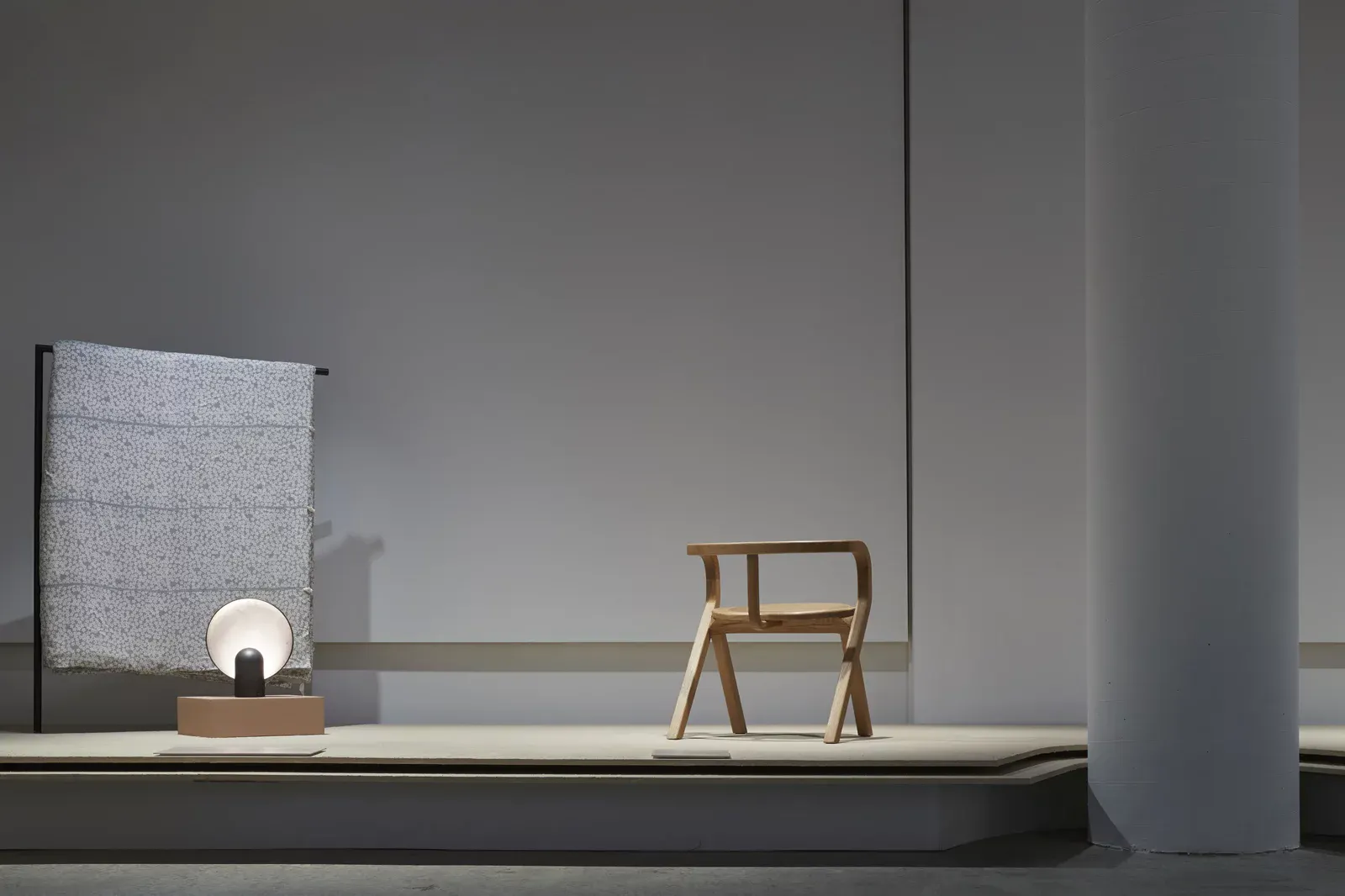 Installation view of Common Good at the Powerhouse Museum. A minimal grey gallery setting displays a small wooden chair maquette and a rounded table lamp on a low plinth, with a textured panel mounted on the wall behind. U-P(co)2010
