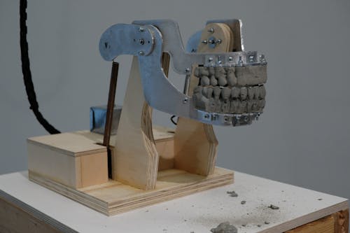 A mechanical sculpture in the shape of a human jaw, made out of aluminium, plywood and cement sits on top of a white pedestal.