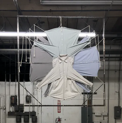 A sculpture hangs suspended from the ceiling. Two invisible wires hold up this hollow square made of steel, one on the top right corner and one on the top left corner. At the center of this hollow square, is 4 long sleeve collared button up work shirts. The necks of these shirts all meet in the center of the hollow square,  and the bottom hem of each shirt points out––the sleeves of the shirts do the same. Each corner of the bottom hem of each shirt and every sleeve has a piece of cotton rope tied to it. This cotton rope is then pulled towards the outer walls of the hollow square, and it is wrapped and tied to the metal bars. Lastly, the cotton rope that remains after being tied dangles down.  