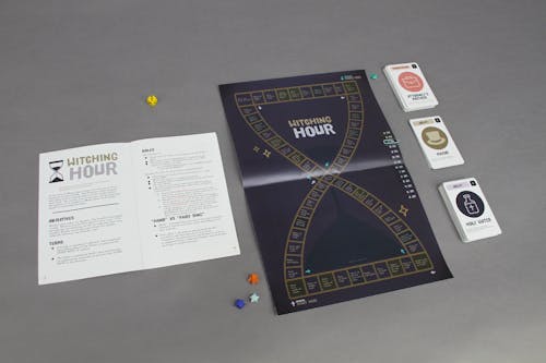 A board game set complete with a board, game pieces, and playable cards in an illustrative style depicting "witchy" motifs such as hourglasses and sparkles.