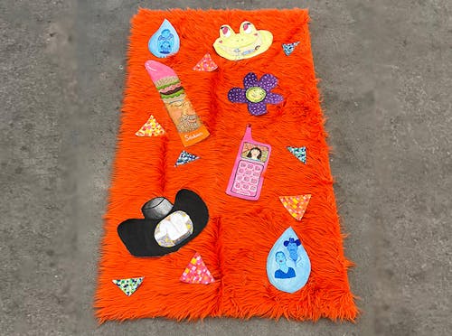 There are 15 small gouache paintings velcroed to a fluffy orange rug. 8 of the paintings are small triangles scattered around the rest of the bigger paintings. The paintings located at the top are a monochrome blue teardrop of two little boys, a frog with pink eyes and its smile has a shower inside. The water for the shower has 3 strings of blue beads to mimic water. Under the teardrop, there is a lipstick-shaped canvas with a sandwich and a portrait of a man. Next to it, there is a purple daisy and the inside circle is green. The green circle of the flower has a portrait of a person. Under the flower there is an old pink phone with the number buttons on it and the small screen has a portrait of a woman. On the bottom left, there is a black cowboy hat and the inside of the hat has a man’s body with a white shirt and blue jeans. On the bottom right, there is another monochrome blue teardrop of a man and a woman. 