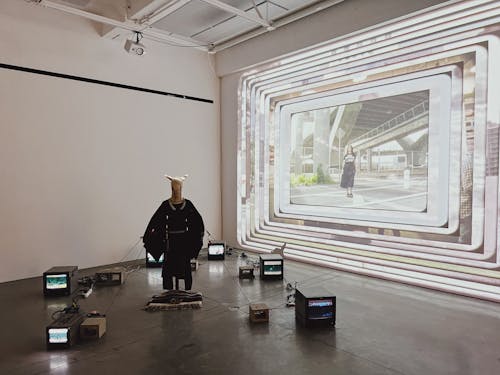 Image of a mannequin wearing a balaclava and a traditional Lakota broadcloth dress connected by wires to six CRTV monitors arranged around the figure in a semi-circle. A large scale video is projected on the wall behind the sculpture.