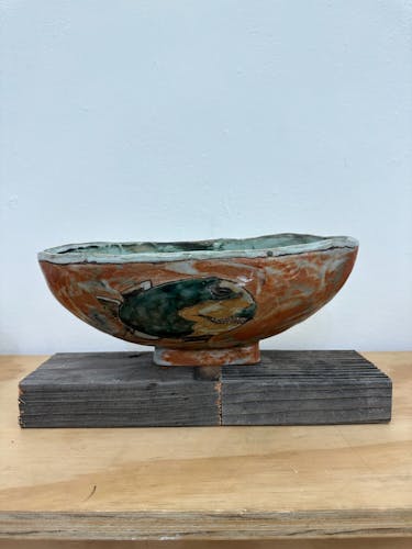 An oblong/oval ceramic bowl or dish about 10 inches wide, 4 inches deep, and 6 inches tall sits atop a gray piece of scrap wood. The bowl is a brushy dark orange, adorned with a cuerda seca drawing of a gray Shark Week blimp. The backside (not pictured) is adorned with another cuerda seca drawing of a white guiding arrow found on a hiking trail that was used for target practice. 