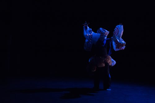 A solo dancer is barely lit in a mostly dark stage. Their arms are outstretched above their head and their body is covered in Target bags. There are bags around this dancer’s waist, arms, and a singular bag on their head as if it has become a mask. There is an intensity emitting from the dancer’s movements, as if the dancer is calling for help from a force we cannot see. Photo by Raphael Arndt features Lingfeng Yi.