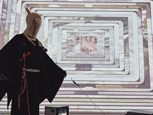 Low angle picture of a mannequin wearing balaclava and a traditional Lakota broadcloth dress positioned in front of a large scale video projection. The video projection has many layers cascading towards the middle where an american flag is seen flowing in the wind.