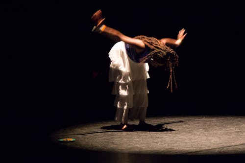A female dancer of African descent is spotlit in a pool of light onstage. She is wearing a metallic blue top and white pants that are layered with ruffles. She is standing with her arms outstretched to the side of her and reaching towards the back. She is bent at the waist with her eyes looking to the ground, as if she is bowing in reverence. To the right of her, there is a small green plate with fried bananas left onstage from a previous section. Photo by Raphael Arndt features Aryeal Lands.