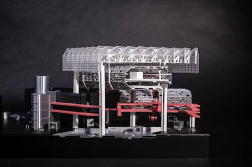 Photograph of a physical model representing a chunk of the entire building proposal. Tall white columns hold up a truss roof structure made of laser cut white acrylic sheets enclosed by an outer shell of frosted translucent acrylic panels. Hovering below the truss system is the community library represented by horizontal hexagonal tube-like structure made of metallic silver 3D prints. A network of catwalk pathways floats in mid-air connecting the different volumes to provide circulation.  