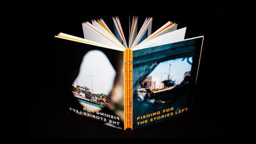 A print book stands in a dark environment, showing its cover and back cover. The cover features a ship peeked through the hole of a canvas wall. The back cover features stacked cargo boxes shot from behind a sculpture.