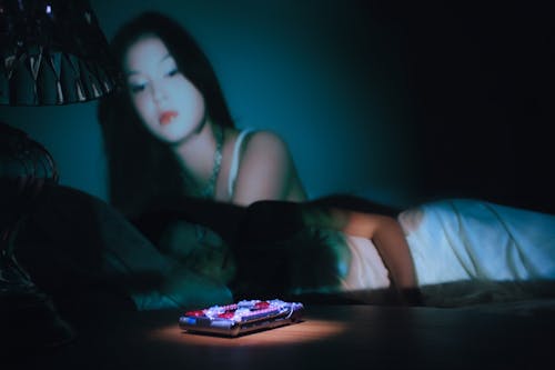 A pink bedazzled Motorola phone is being spotlit as a girl in a white night gown is sleeping on a bed. A projection of her ghost is seen watching over her.