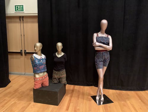 Two half-body mannequins on a shared podium and one full body mannequin on the right. Each have different outfits on. The mannequin farthest left wears a blue Peter Pan collar top with orange shorts. The mannequin in the middle wears a black v-neck t-shirt and black and brown striped shorts. The third mannequin wears a blue romper with zizag patterns. It also has red straps and a matching red belt.