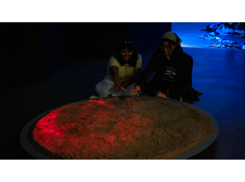 Two women sitting at a round platform. The surface is covered in sand and their hands hover on top. The background is dark with a blue glow. There is a red projection of grunion fish onto the sand. 