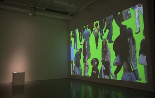 large white gallery space, large full wall projection of neon green background with figures of individual people having blurred faces scattered throughout the image and posed in walking motions.