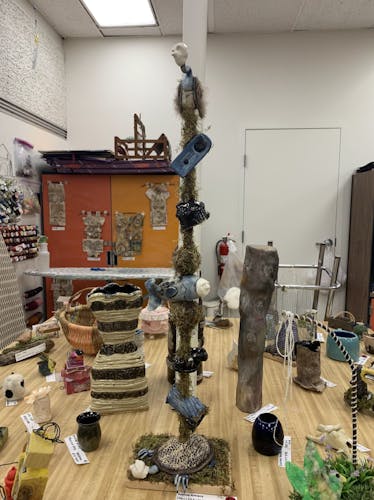 Large totem pole with creepy blue animal figures (vulture, fish, turtle) wearing human masks and various objects, in order: vulture, grandfather clock, hut, moss ball, fish, moss ball, mushrooms, tree stump, turtle with hands, moss covered base